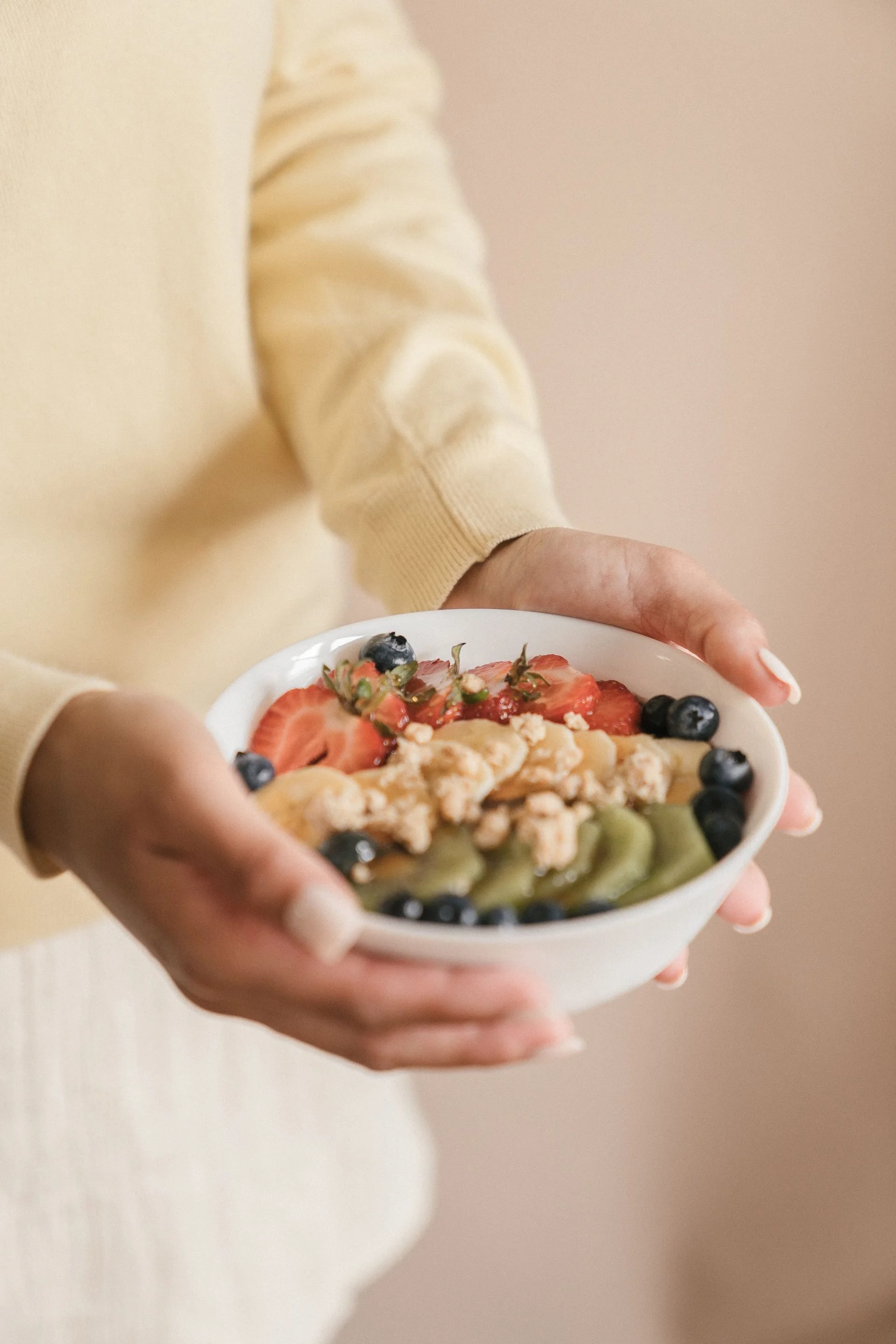 Person holding a bowl of mixed fruit, including strawberries and blueberries, against a beige background.