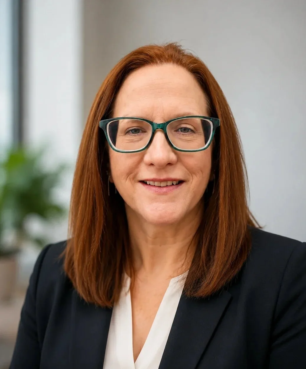 Rebecca Ferrell, a woman with shoulder-length red hair and glasses, wearing a black blazer and white blouse, smiling in an indoor office setting.