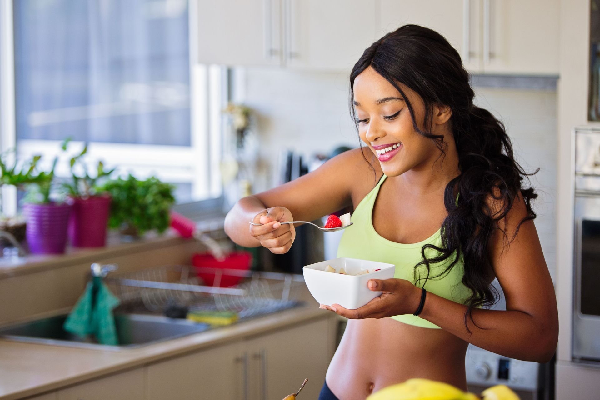 A young woman with long dark hair, wearing a yellow sports bra, smiling while holding a spoon with strawberries and yogurt in a kitchen.
