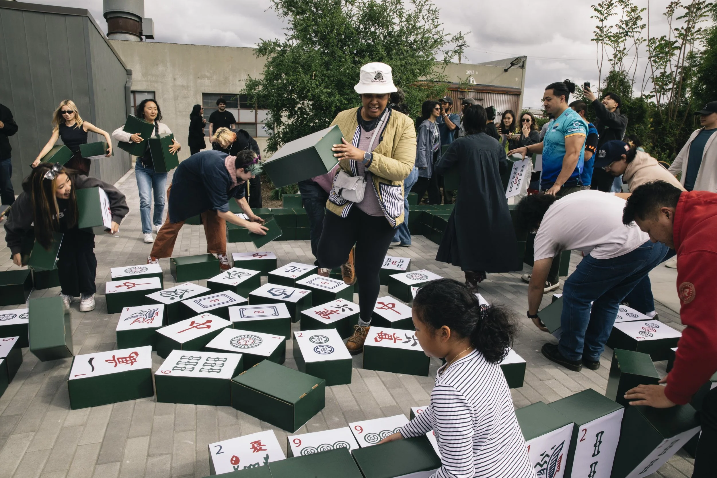 A large group of people playing a game outdoors, with Mahjong tiles arranged on the ground. Some people are holding boxes of Mahjong tiles, and others are gathered around, actively participating or observing. The setting appears to be a patio or outd