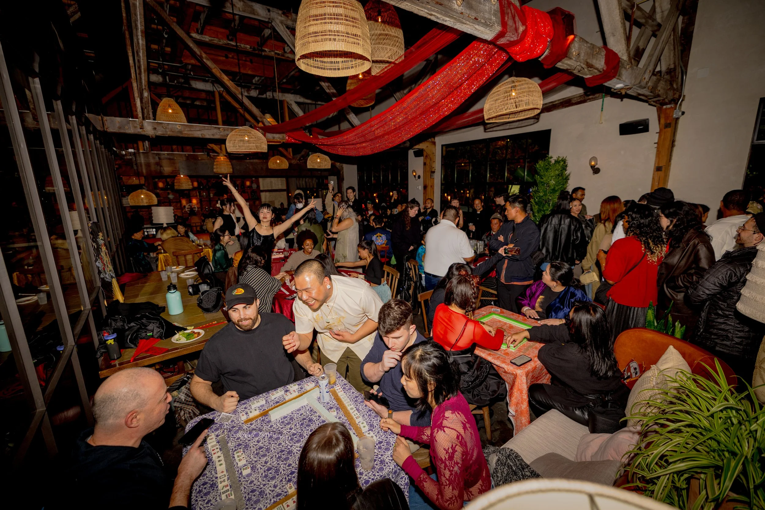 A large indoor gathering of diverse people engaged in socializing and playing games, with decorated ceiling featuring hanging woven lamps and red fabric draped across the room.