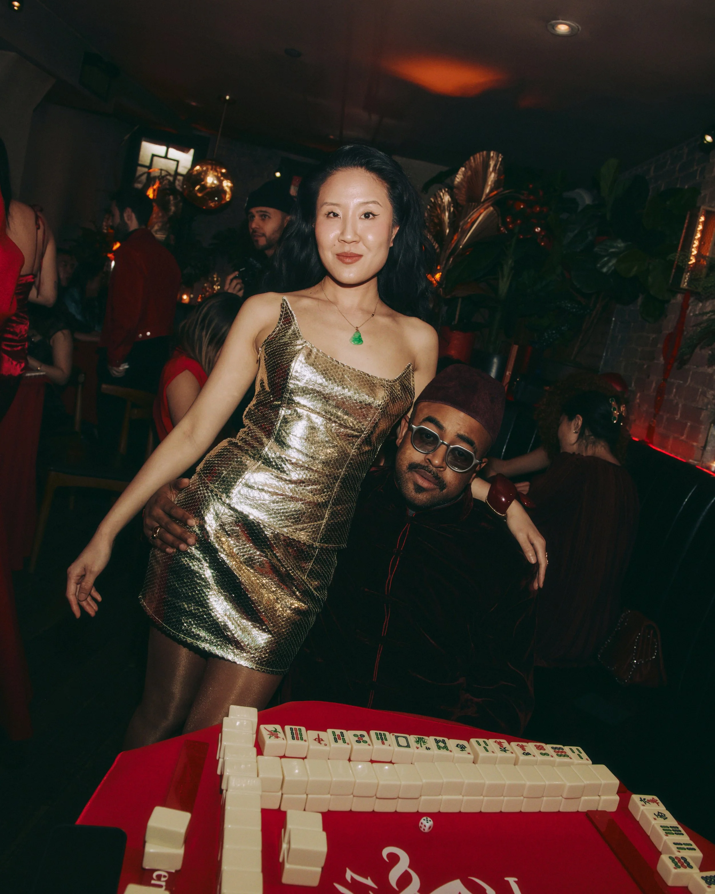 A woman in a gold dress stands next to a man with glasses at an indoor party, with a mahjong game on a table in the foreground, and other guests in the background