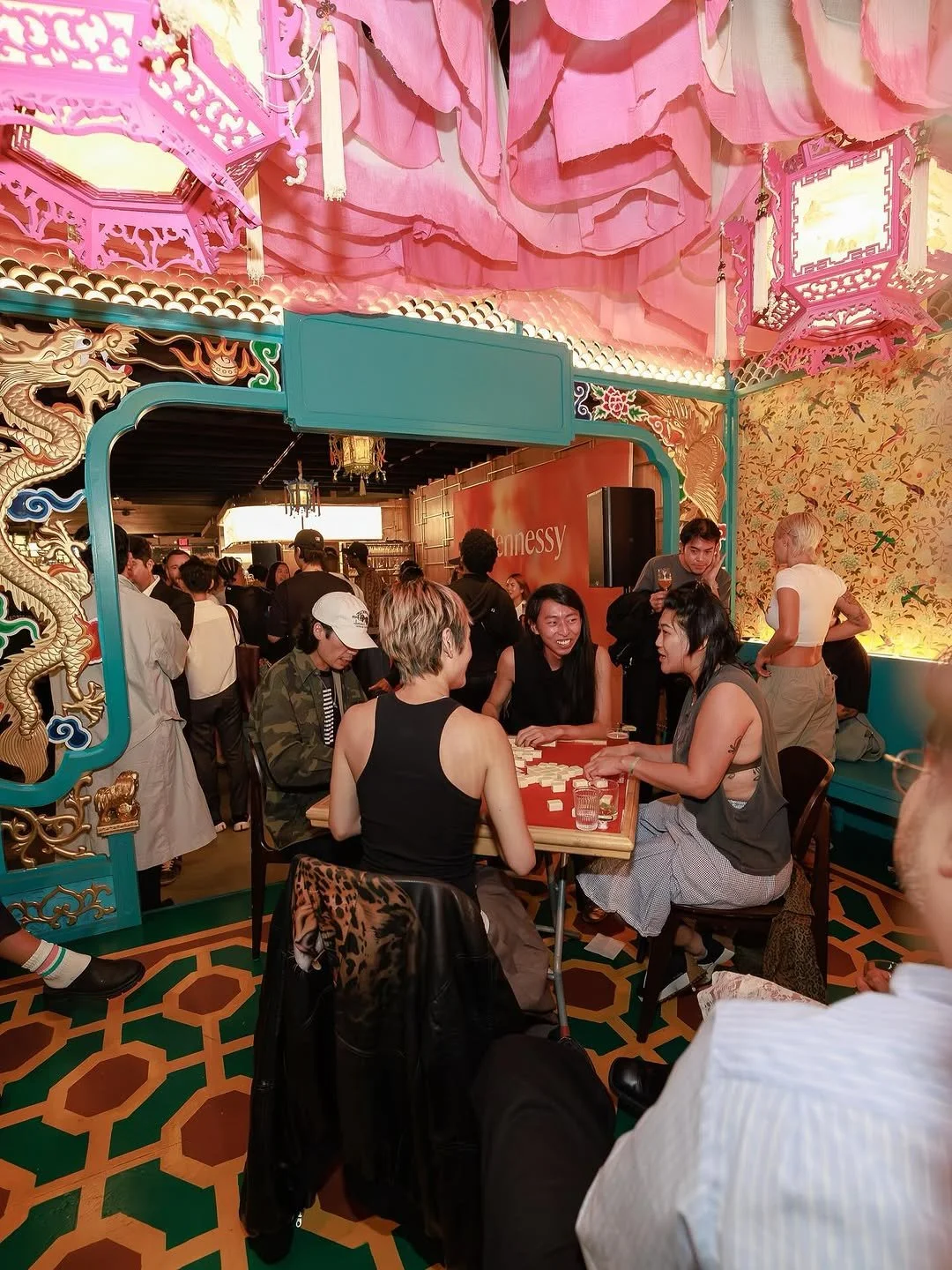 People playing mahjong at a table in a lively Asian-themed restaurant with dragon and floral decorations, colorful walls, and pink fabric ceiling drapes.