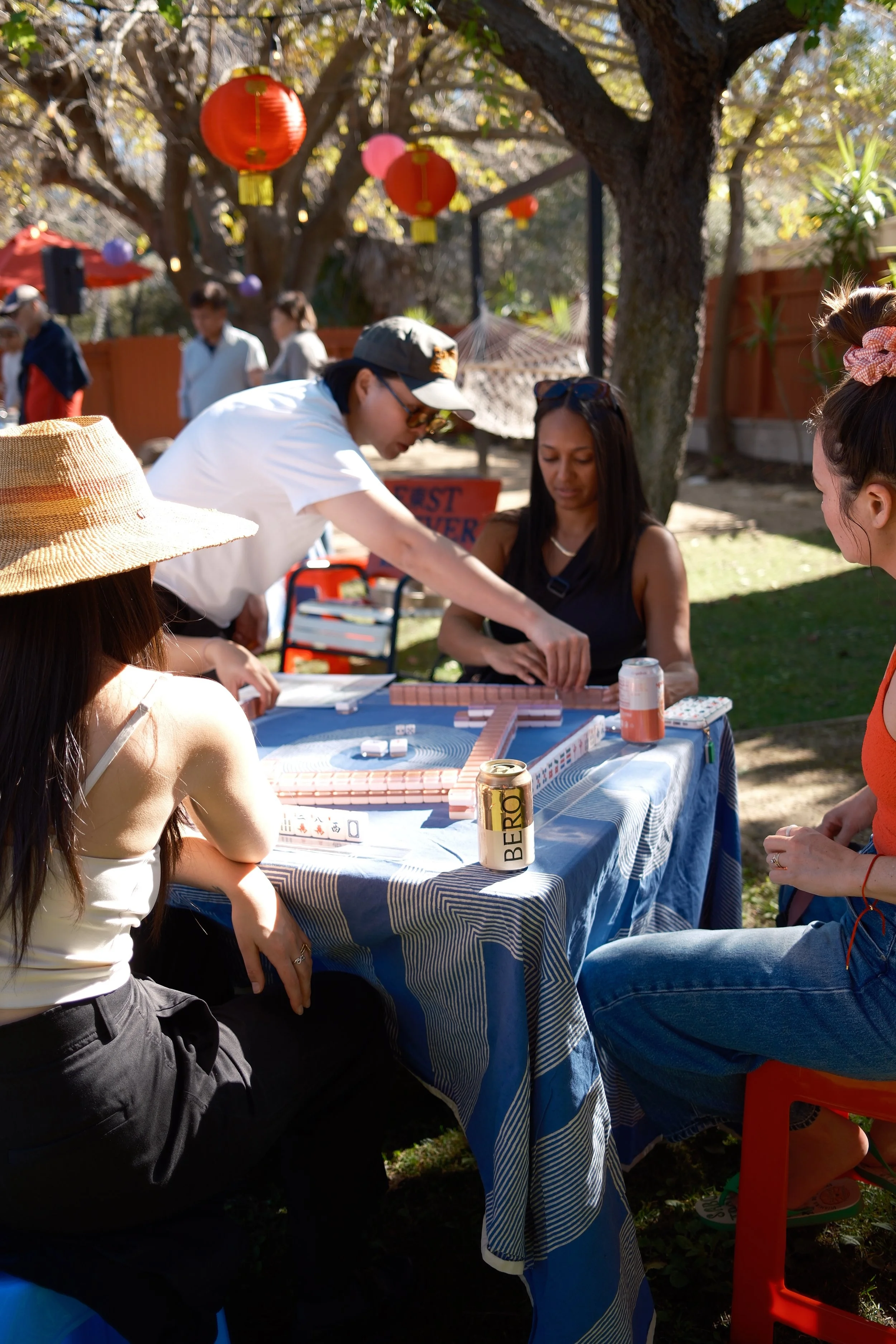 People gathered outdoors around a table playing Mahjong during the daytime. There is a tree with hanging lanterns in the background.