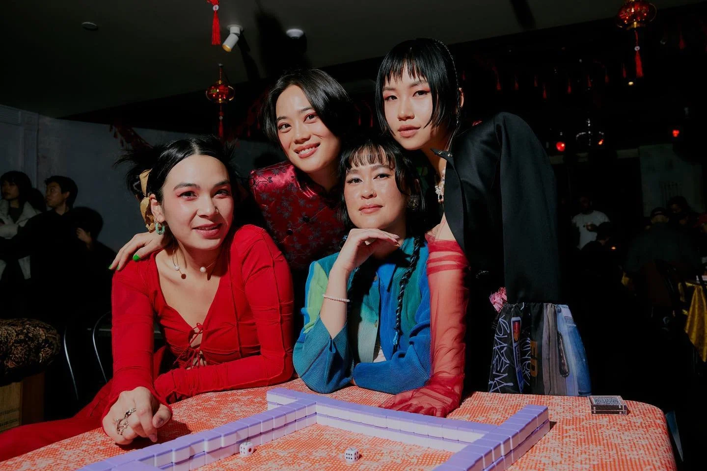 Four women around a table playing a game with dominoes and dice, smiling and posing for a photo in a dimly lit restaurant or event space, with decorations hanging from the ceiling.