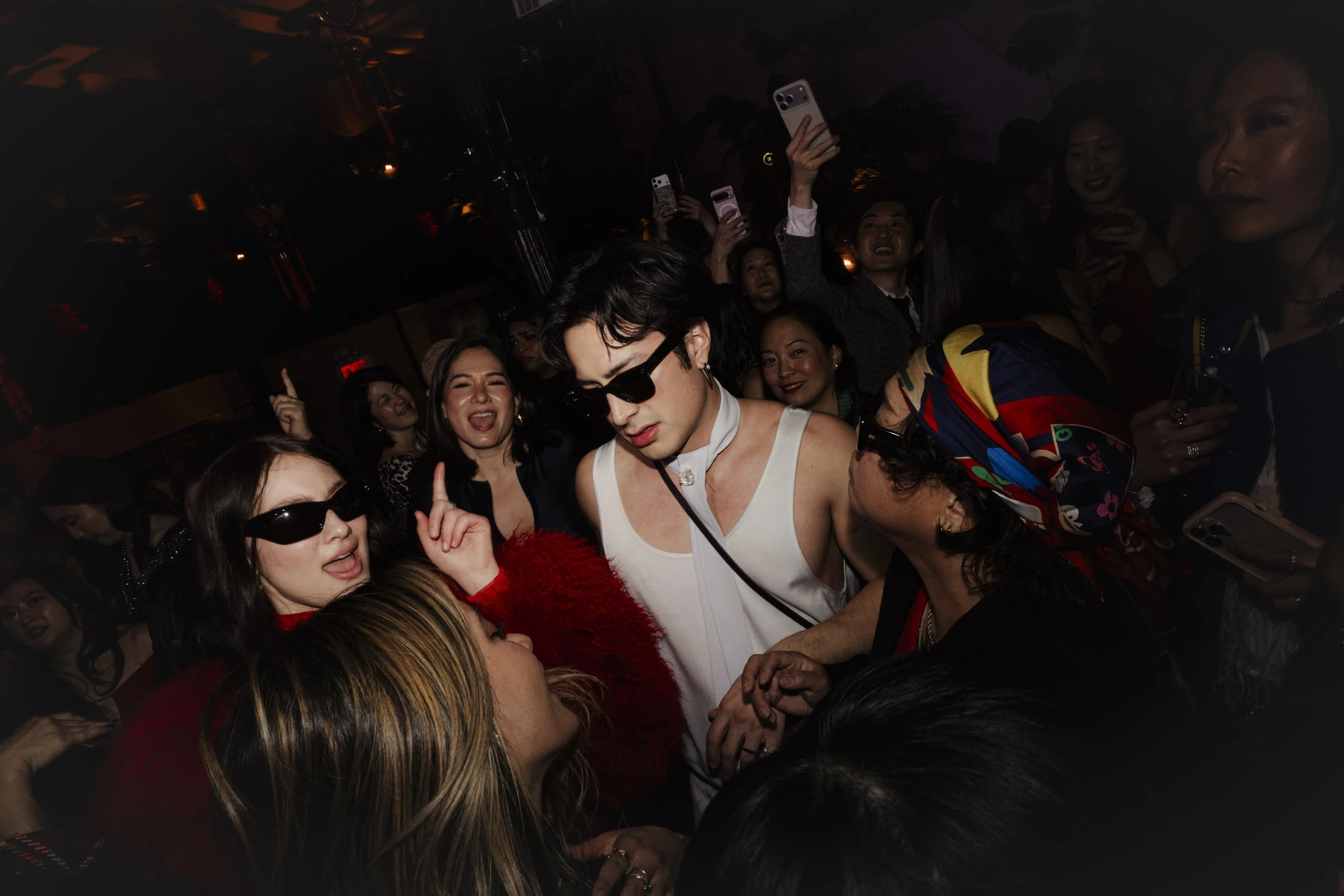 A crowded indoor gathering with people taking photos and dancing at a party or celebration, with dim lighting and vibrant atmosphere.
