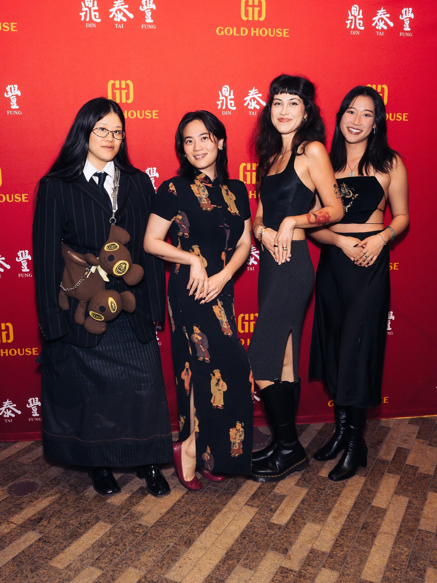 Four women standing in front of a red background with yellow and white text and logos. They are dressed in fashionable clothing, with one wearing a pinstripe suit, another in a dress with traditional Asian patterns, a third in a black dress with flor