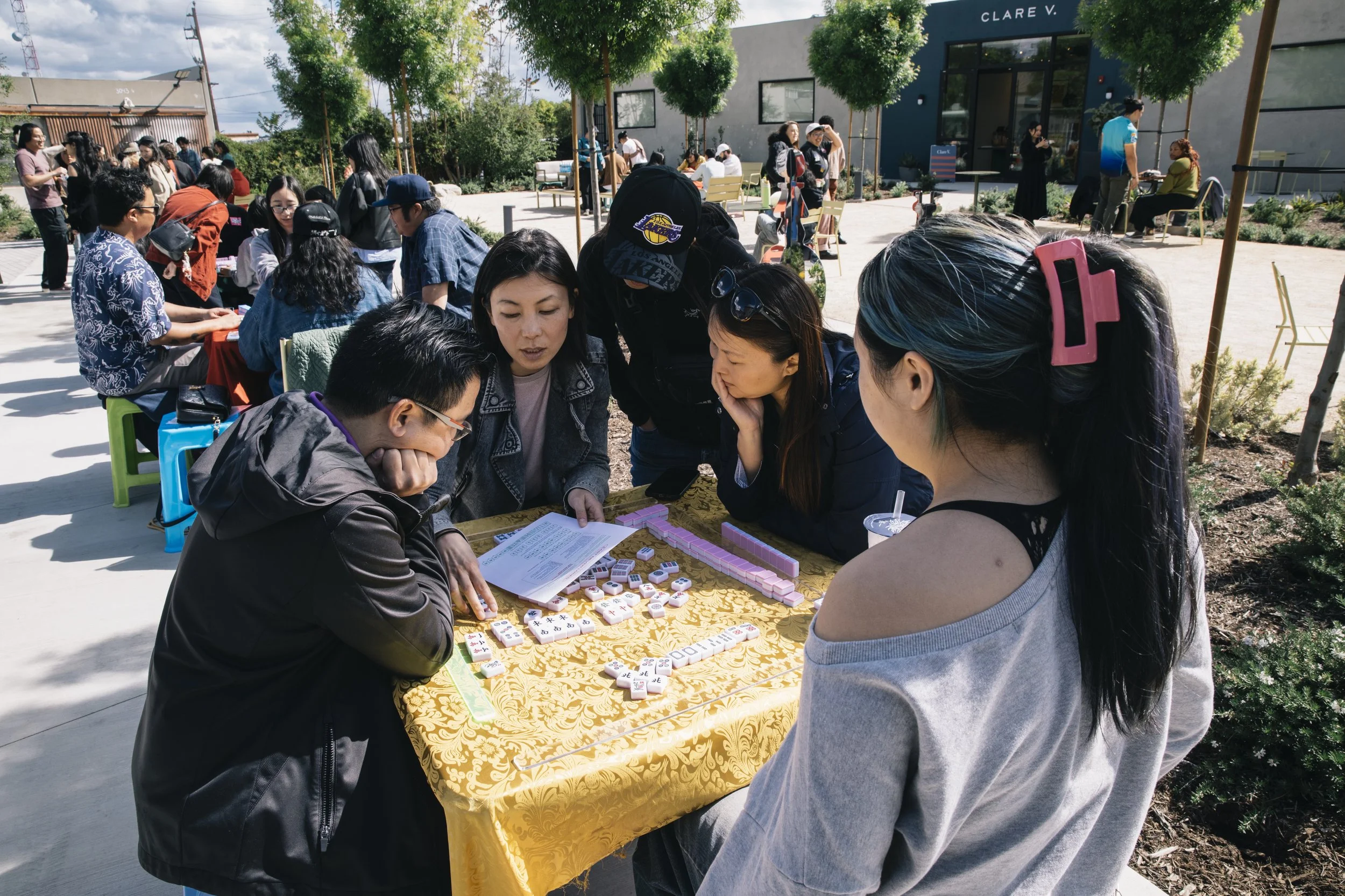 People gathered outdoors playing mahjong at a long table covered with a yellow tablecloth. Some are focused on the game, while others are observing or chatting. There are trees and a building in the background.