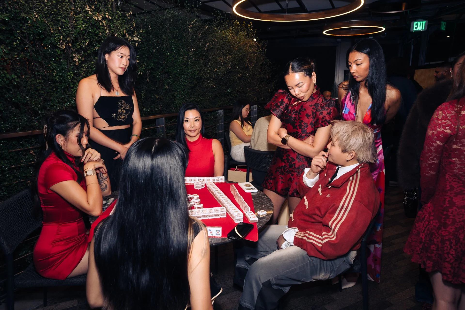 Group of women and a man playing Mahjong at a table in a restaurant or event space with a green wall and modern lighting