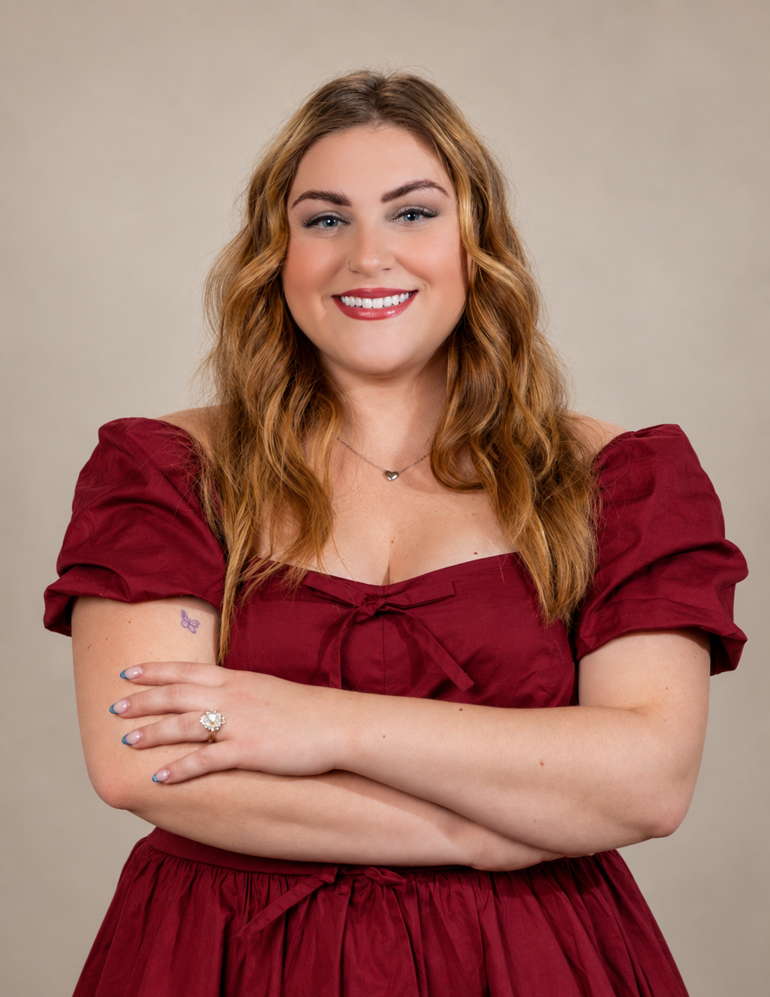 A woman with wavy red hair and fair skin smiling, wearing a red dress with puffed sleeves and a bow at the neckline, standing against a beige background.