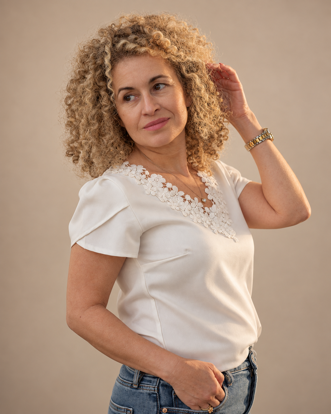 A woman with curly blonde hair, wearing a white blouse with floral embroidery around the neckline, and blue jeans, posing against a plain beige background.