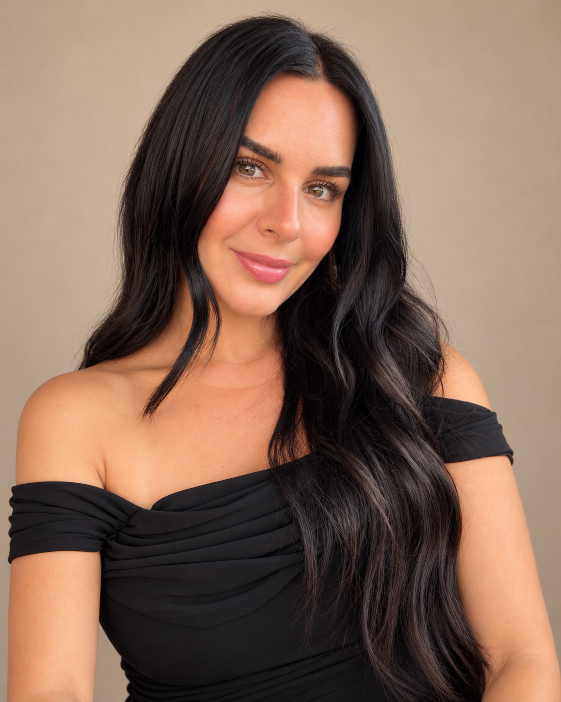 A woman with long, wavy black hair and light skin, wearing a black off-the-shoulder top, smiling softly against a beige background.