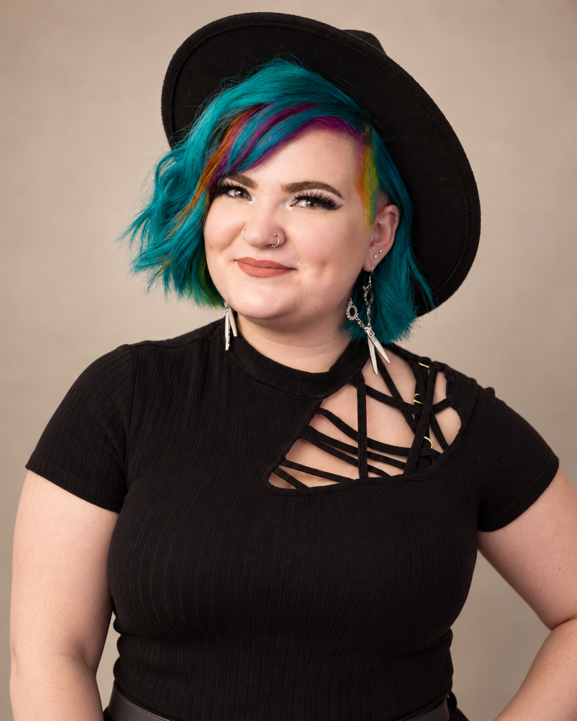 A young woman with brightly colored blue, purple, and yellow hair, wearing a black hat, a black top with crisscross details on the chest, and dangling earrings, smiling at the camera.