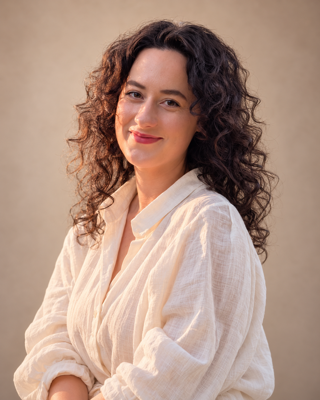 A woman with curly dark hair wearing a light-colored button-up shirt, smiling and looking at the camera with her arms crossed.