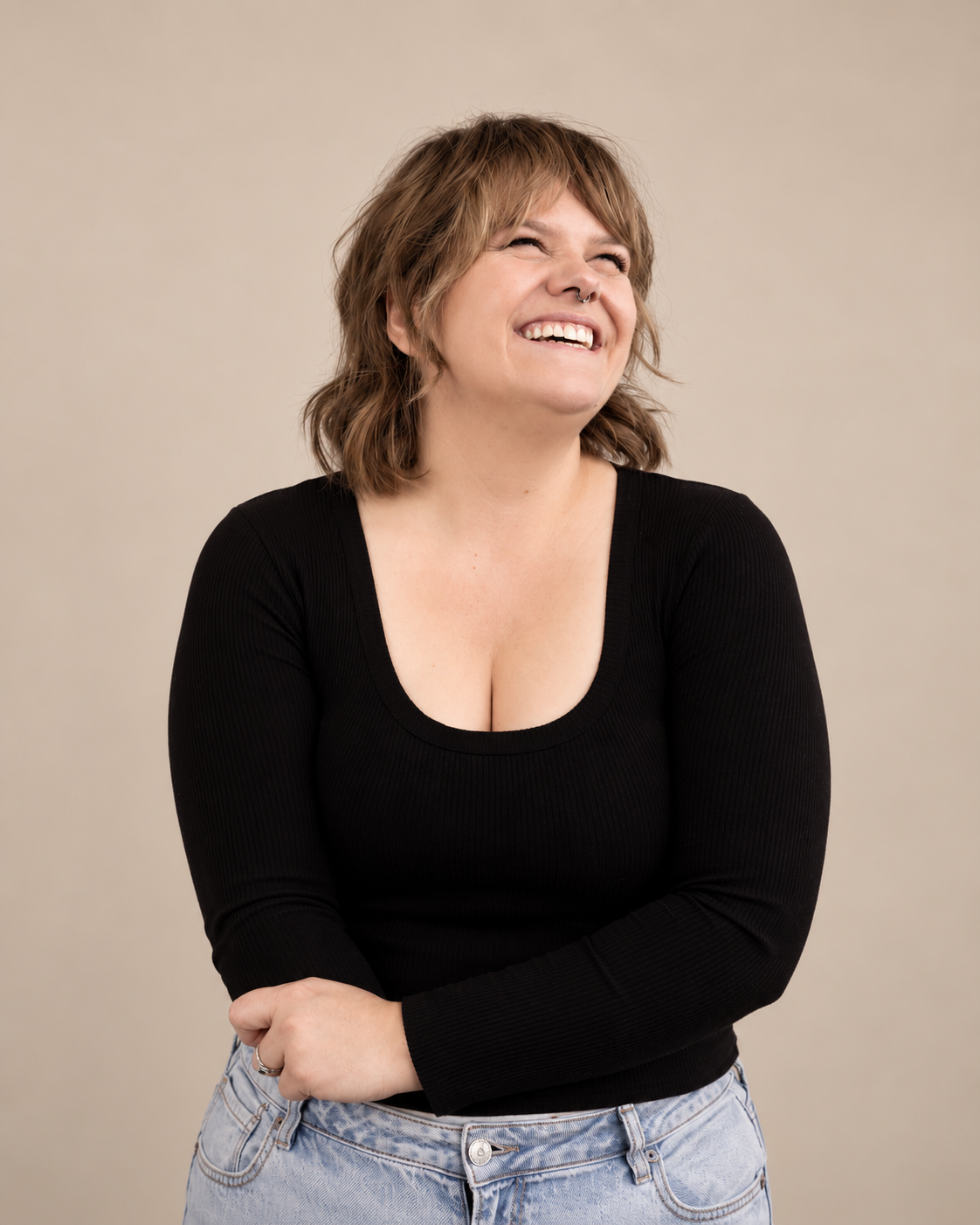 A woman with short, wavy brown hair smiling and laughing, wearing a black long-sleeve top and light blue jeans, standing against a plain beige background.