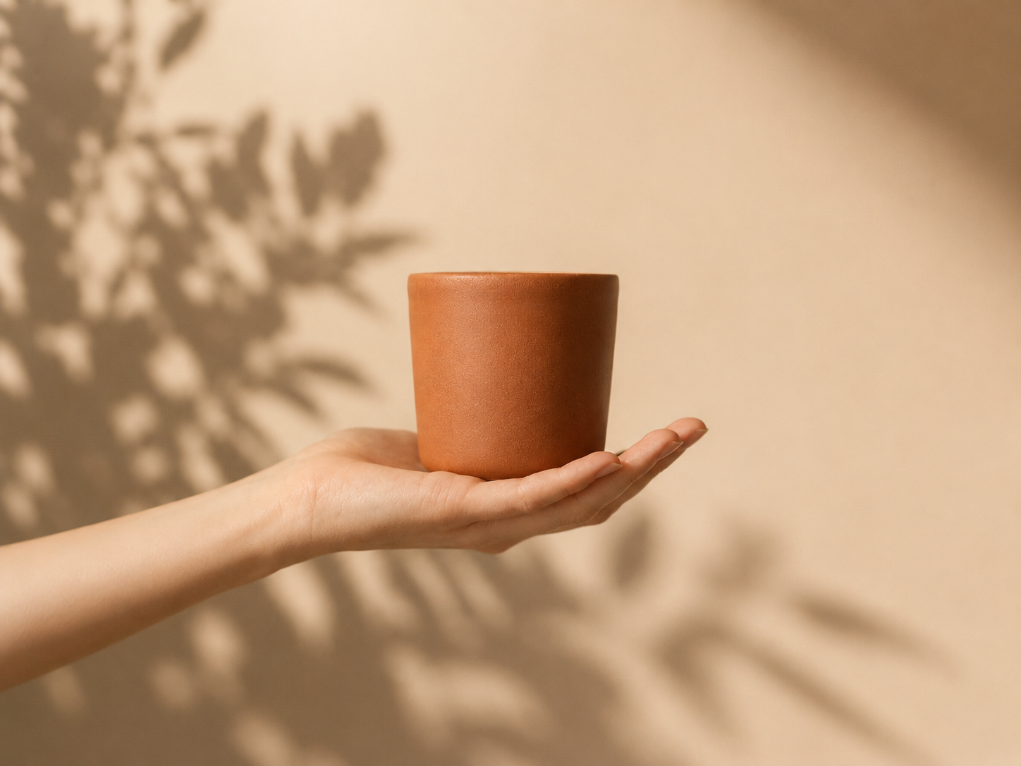 A person's hand holding a small, empty terracotta plant pot against a beige background with shadow of leaves.