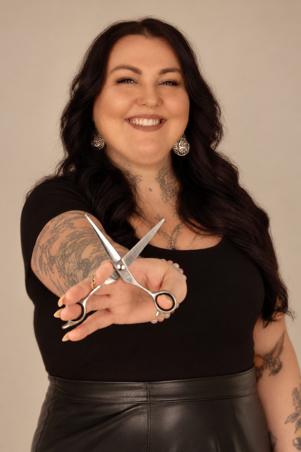 A smiling woman with long dark hair, tattoos, earrings, and a nose piercing, holding a pair of scissors directed towards the camera.