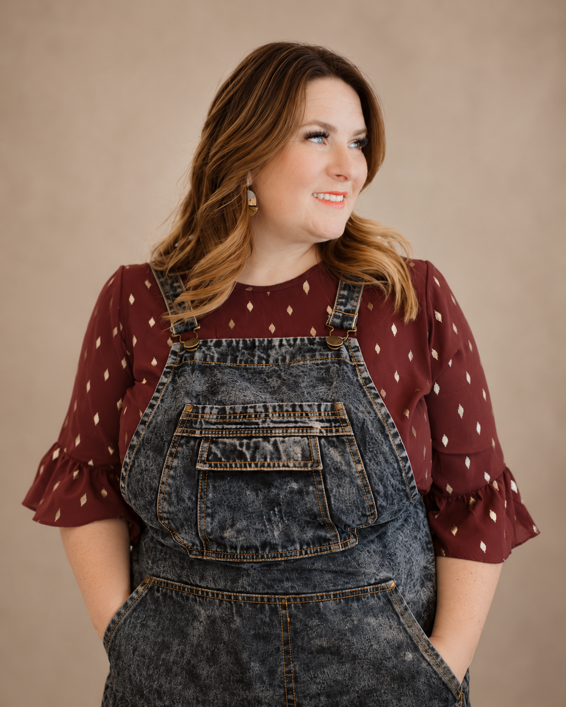 A woman with shoulder-length red hair, wearing a maroon blouse with ruffled sleeves and a diamond pattern, and acid-wash denim overalls, smiling and looking to her right against a plain beige background.