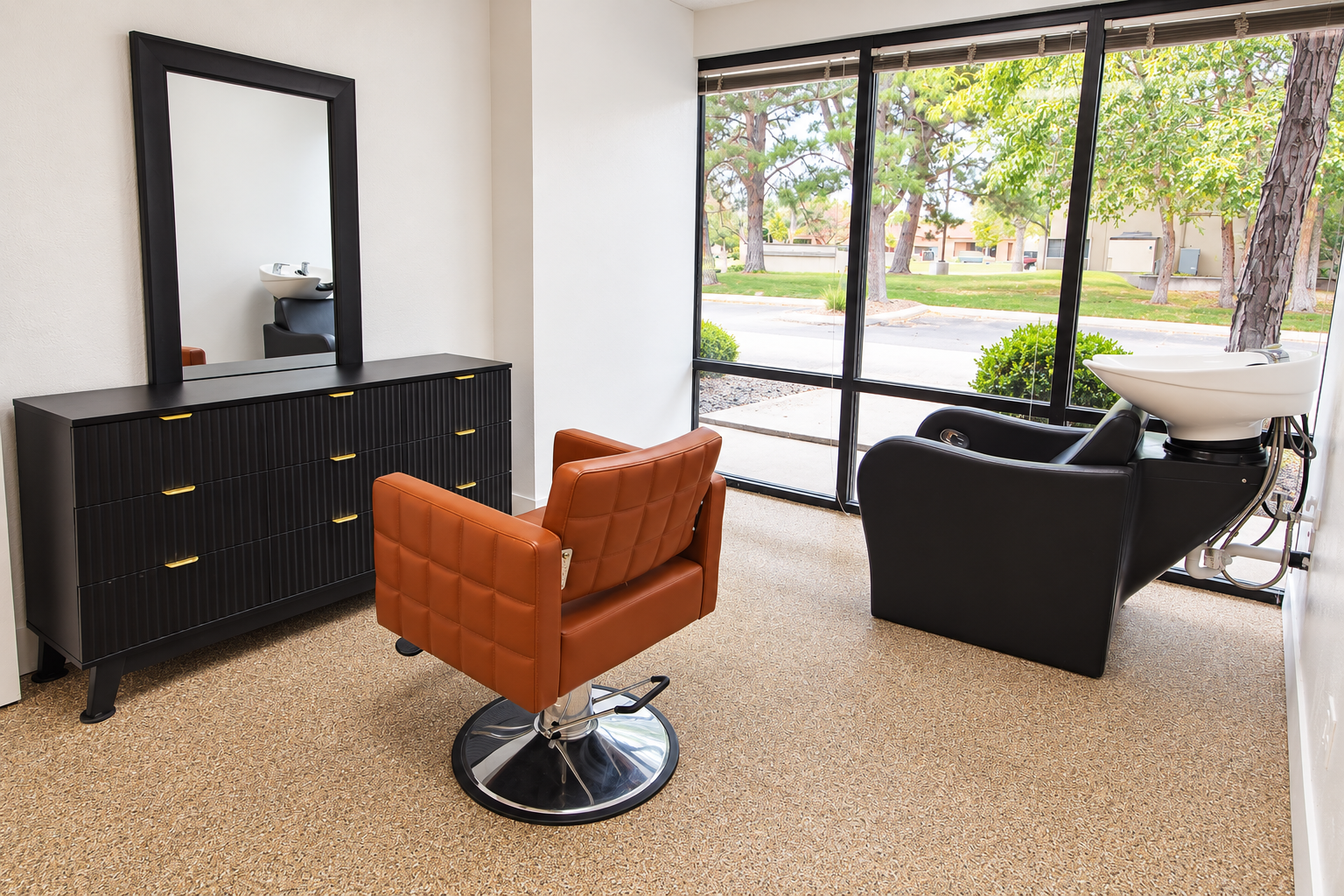 A beauty salon or barber shop interior with a hair-washing sink, a black salon chair, an orange salon chair, a black cabinet with a mirror, and large windows showing trees and a street outside.