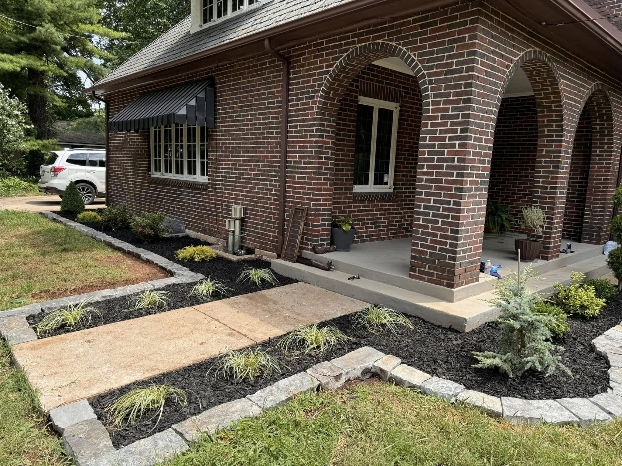 A brick house with arched columns on the porch, window with an awning, a white SUV parked in the driveway, and a landscaped yard with small plants and gravel.