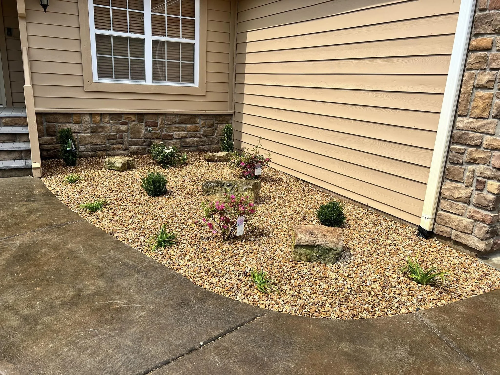 Front yard area with beige siding, a window, potted plants, and a small landscaped garden with rocks and flowers next to the house.