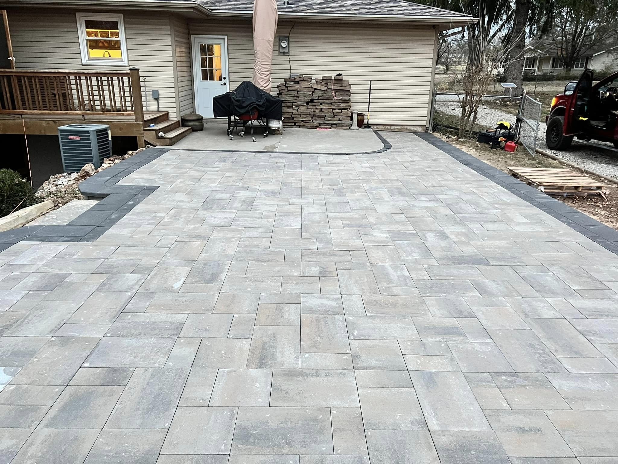 View of a backyard patio with a newly installed patterned stone surface, a door leading inside a house, a small deck with stairs, construction materials including bricks and pallets, and trees in the background.