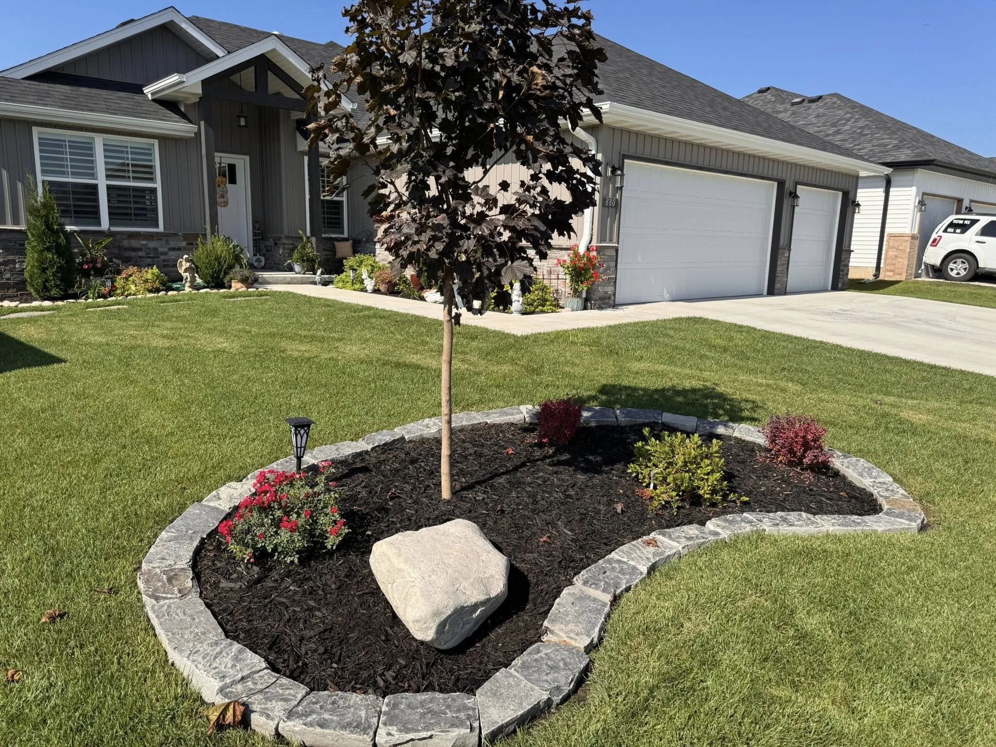 A well-kept front yard with green grass, small trees, and flower beds. A charcoal-colored house with white trim has a two-car garage and a pathway leading to the front door. Potted plants and decorative stones decorate the entrance area. The sky is c