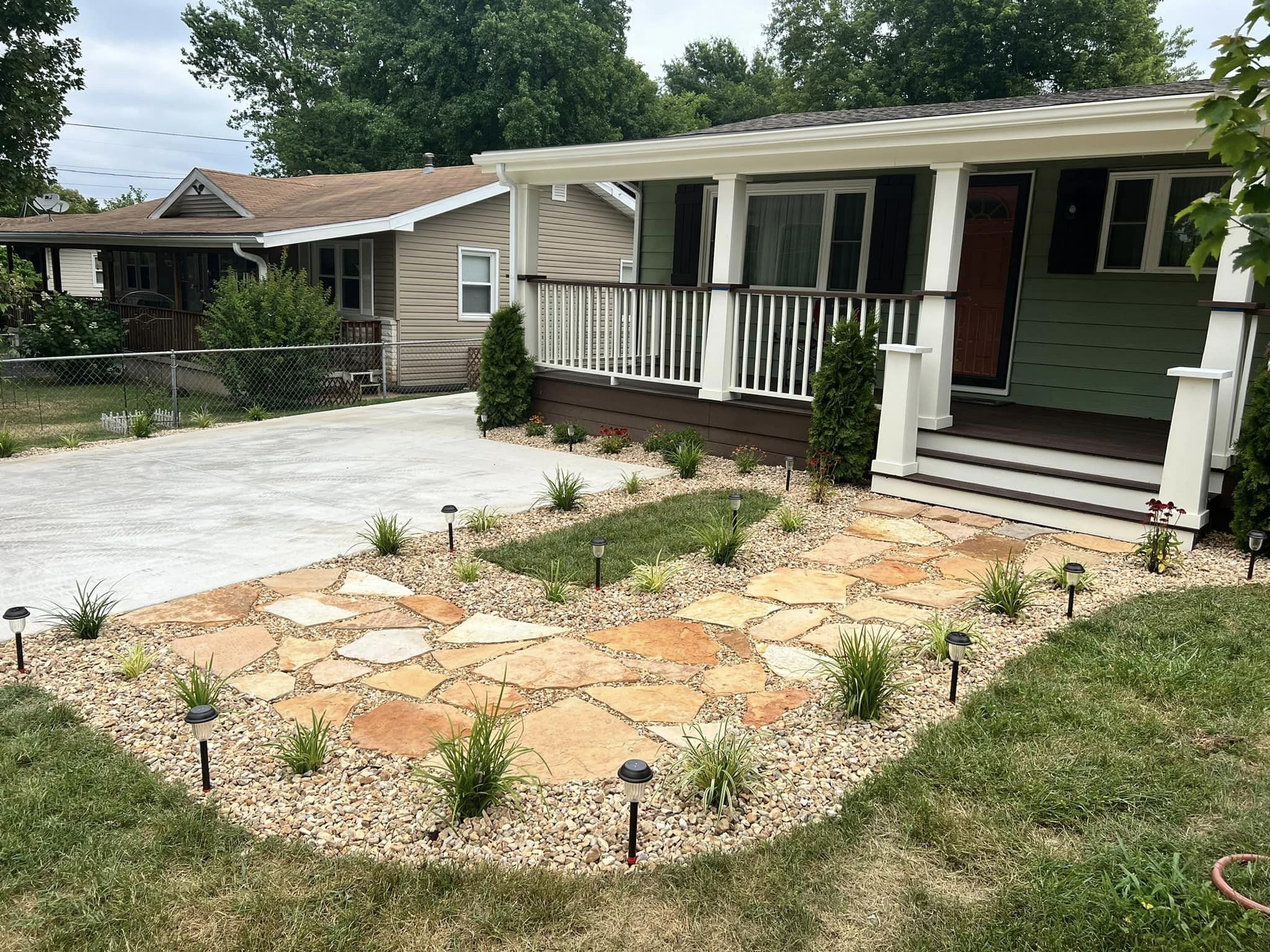 Yard with a circular stone pathway, small plants, grass, and outdoor lighting, with two houses in the background, one with a porch and railing, and trees and cloudy sky overhead.