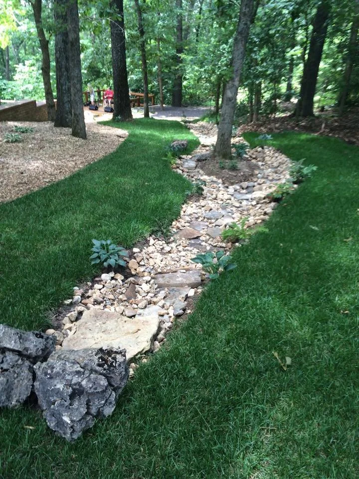 A landscaped yard with a narrow dry creek bed made of rocks and stones, bordered by lush green grass and mature trees in the background.
