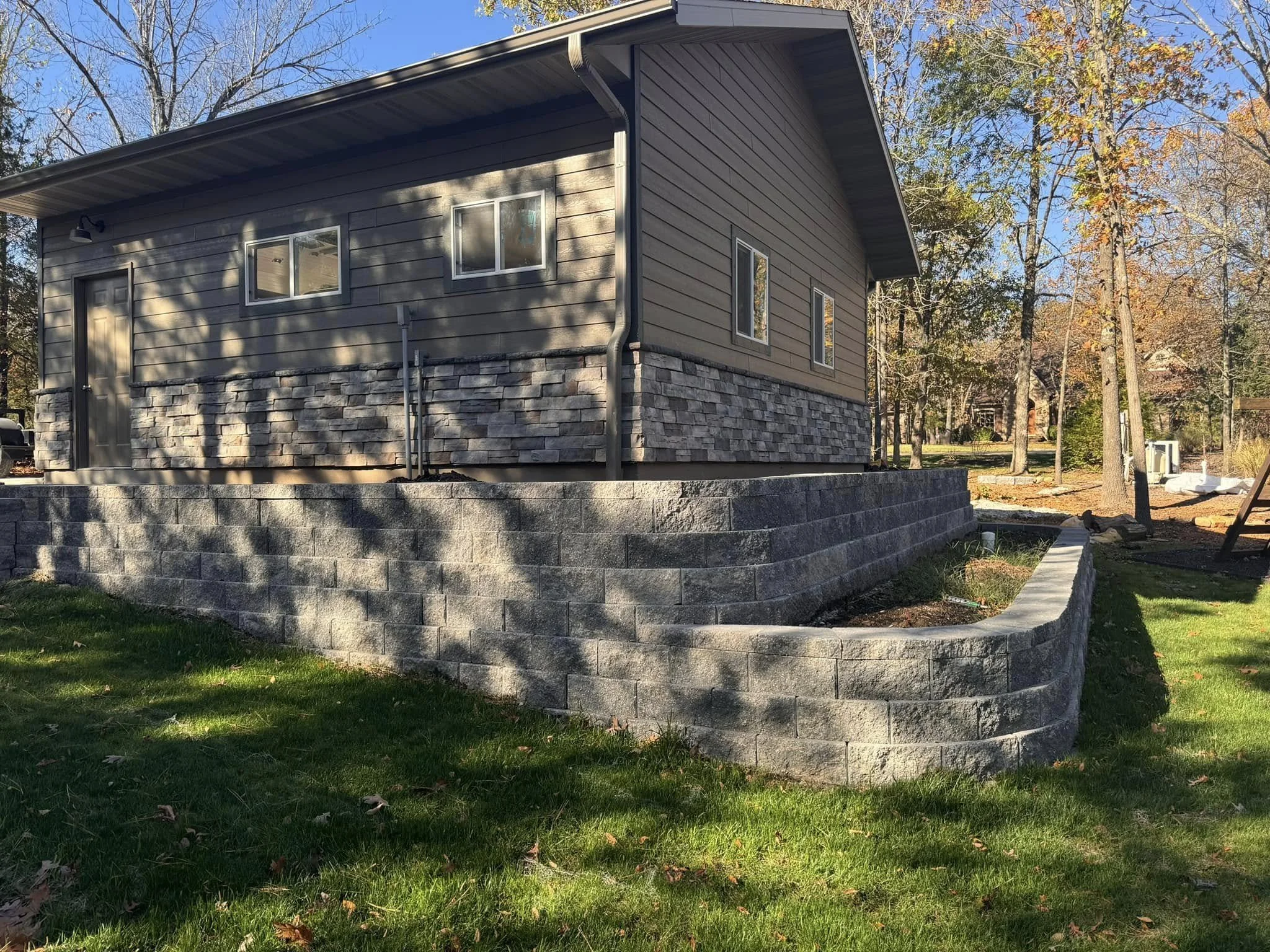 A house with beige siding and a stone foundation, surrounded by trees with fall foliage, on a clear and sunny day.