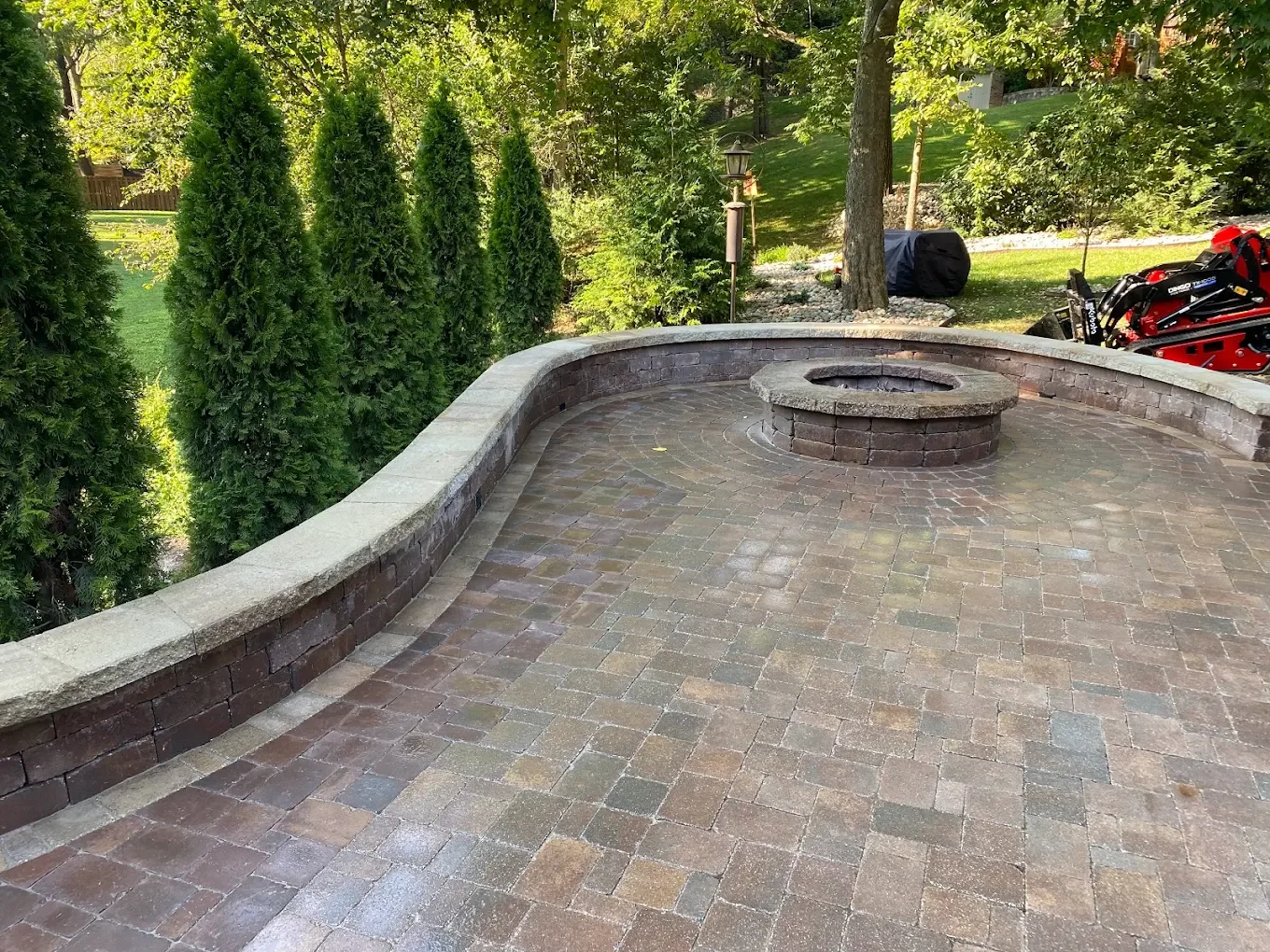 backyard patio with brick paver flooring, a curved brick wall, a fire pit in the center, tall green trees and shrubs, outdoor lighting, and a yard with grass, trees, and a lawnmower in the background