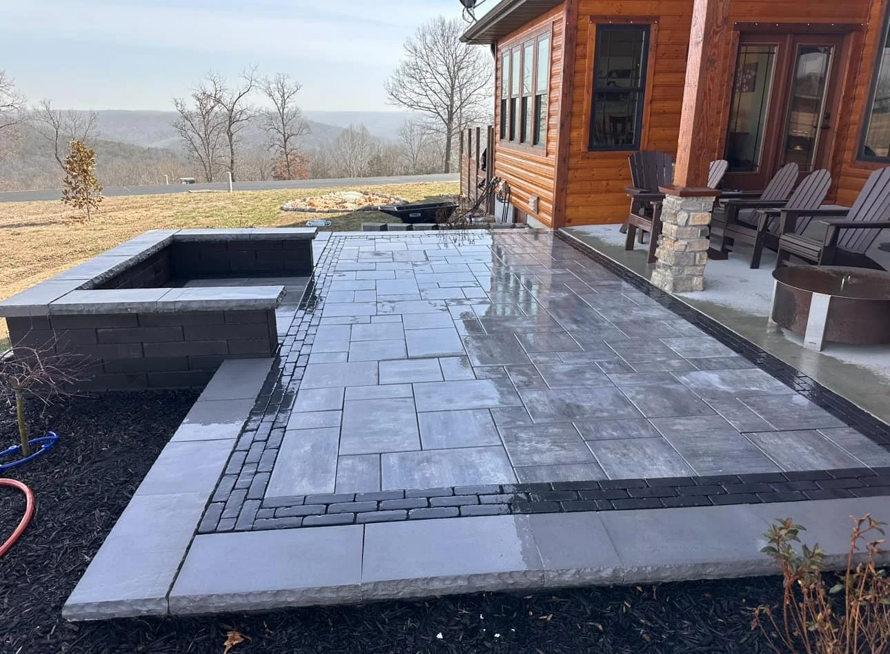 A newly paved stone patio with a built-in fire pit, overlooking a mountainous landscape with leafless trees, attached to a wooden house with outdoor seating and a fire pit.