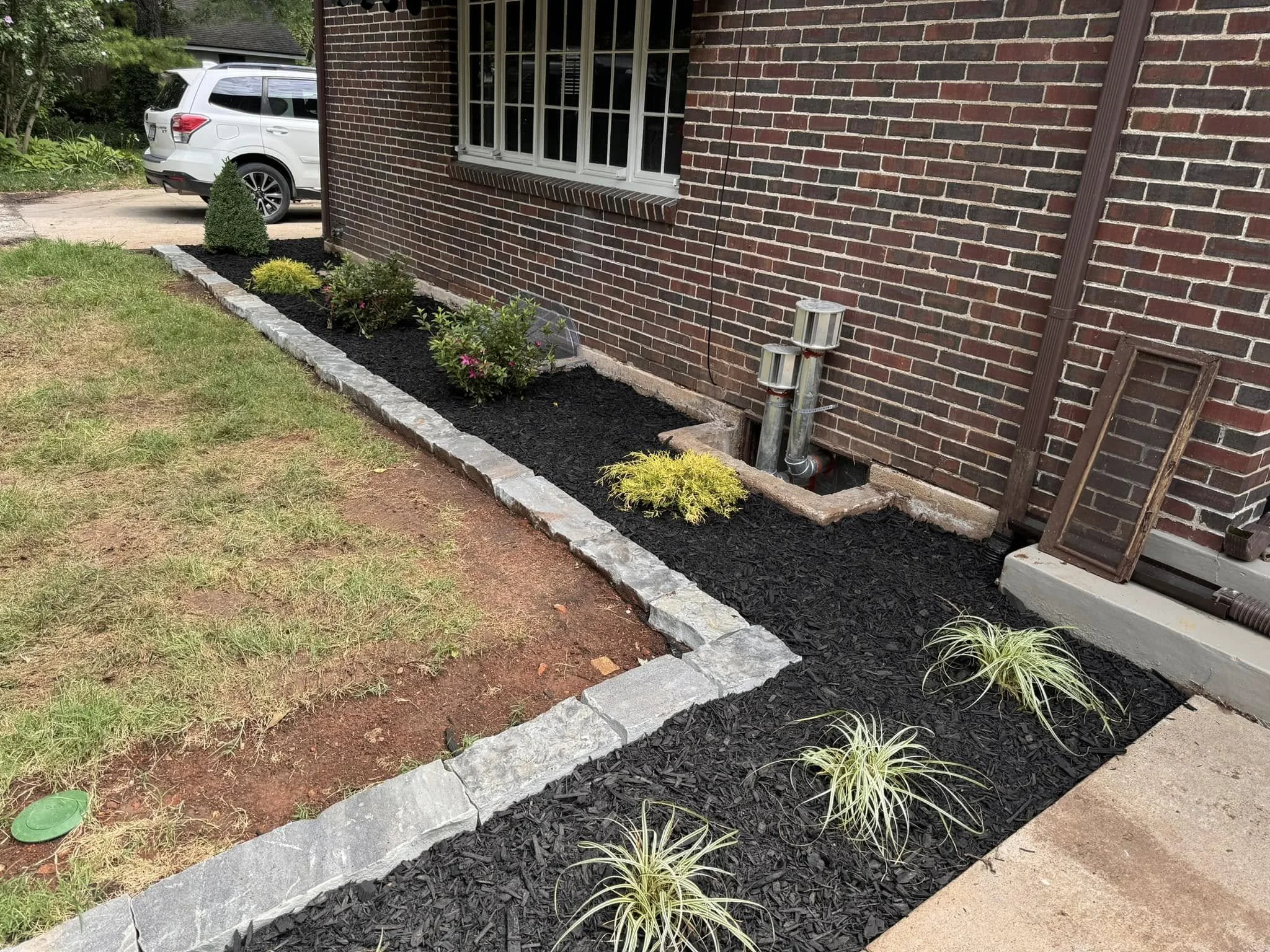 A brick house with a small garden bed along the wall, featuring a small tree, bushes, and ornamental grasses. A white SUV is parked in the driveway in the background.