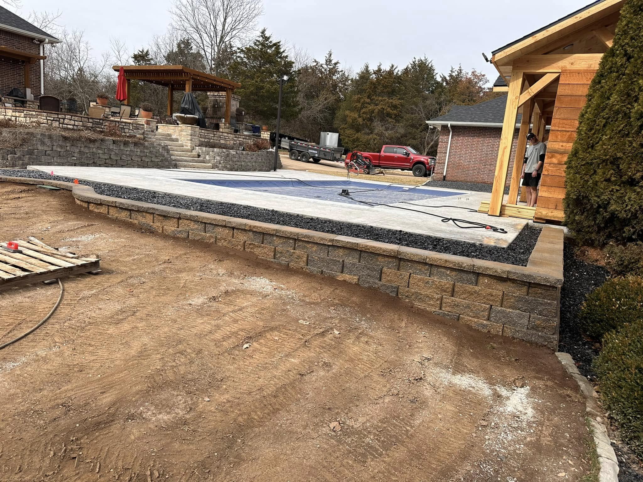 A backyard with under-construction patio area, a raised stone terrace with outdoor furniture and a grill, and a concrete area with a pool cover. There are two houses, one with brick and one with wooden siding, and a worker is standing under a wooden 