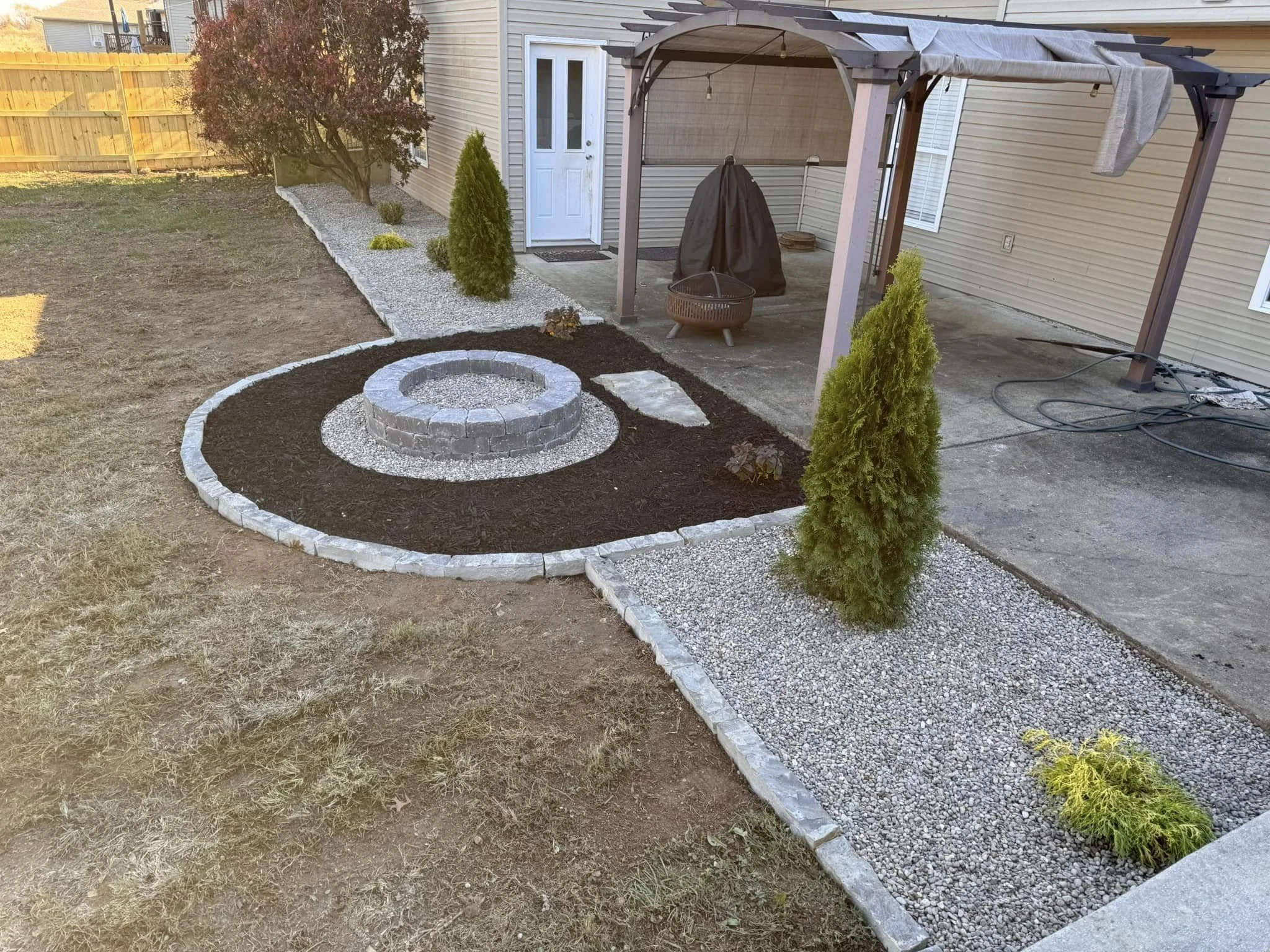 Backyard garden with a circular brick fire pit, newly planted shrubs, and a small gravel path near a house with a pergola.