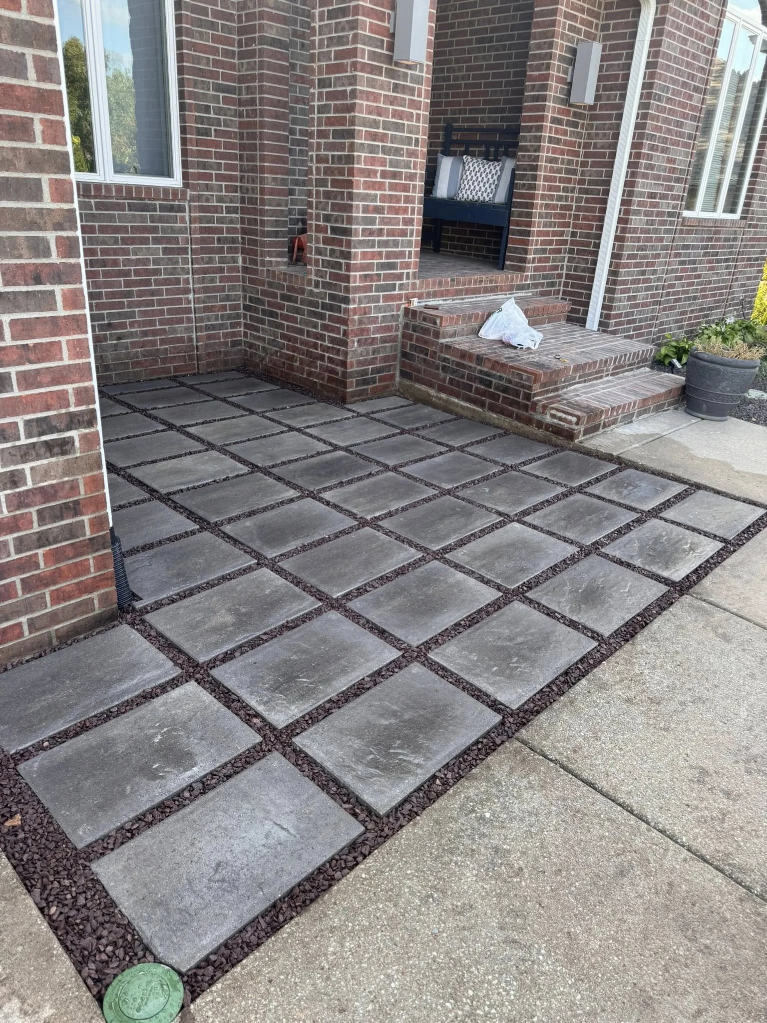 A patio with concrete pavers separated by gravel, brick stairs leading to a porch, brick house walls with windows, and a potted plant.
