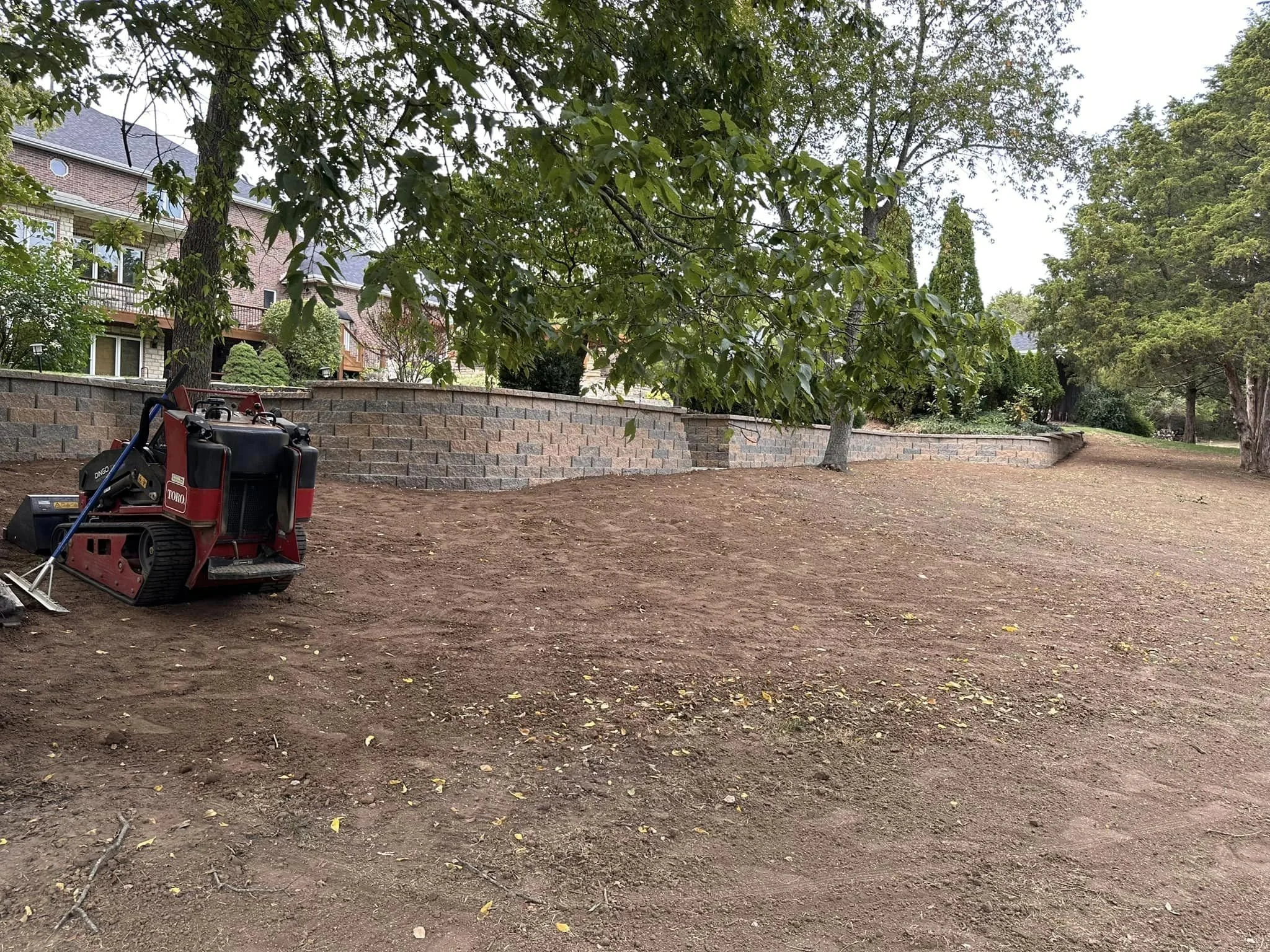 Clean earth with a brick retaining wall, several trees, and house in the background. There is construction equipment in the foreground.