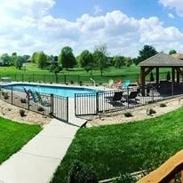 View of a community swimming pool with lounge chairs, surrounded by a black fence and a grassy area, under a blue sky with clouds.