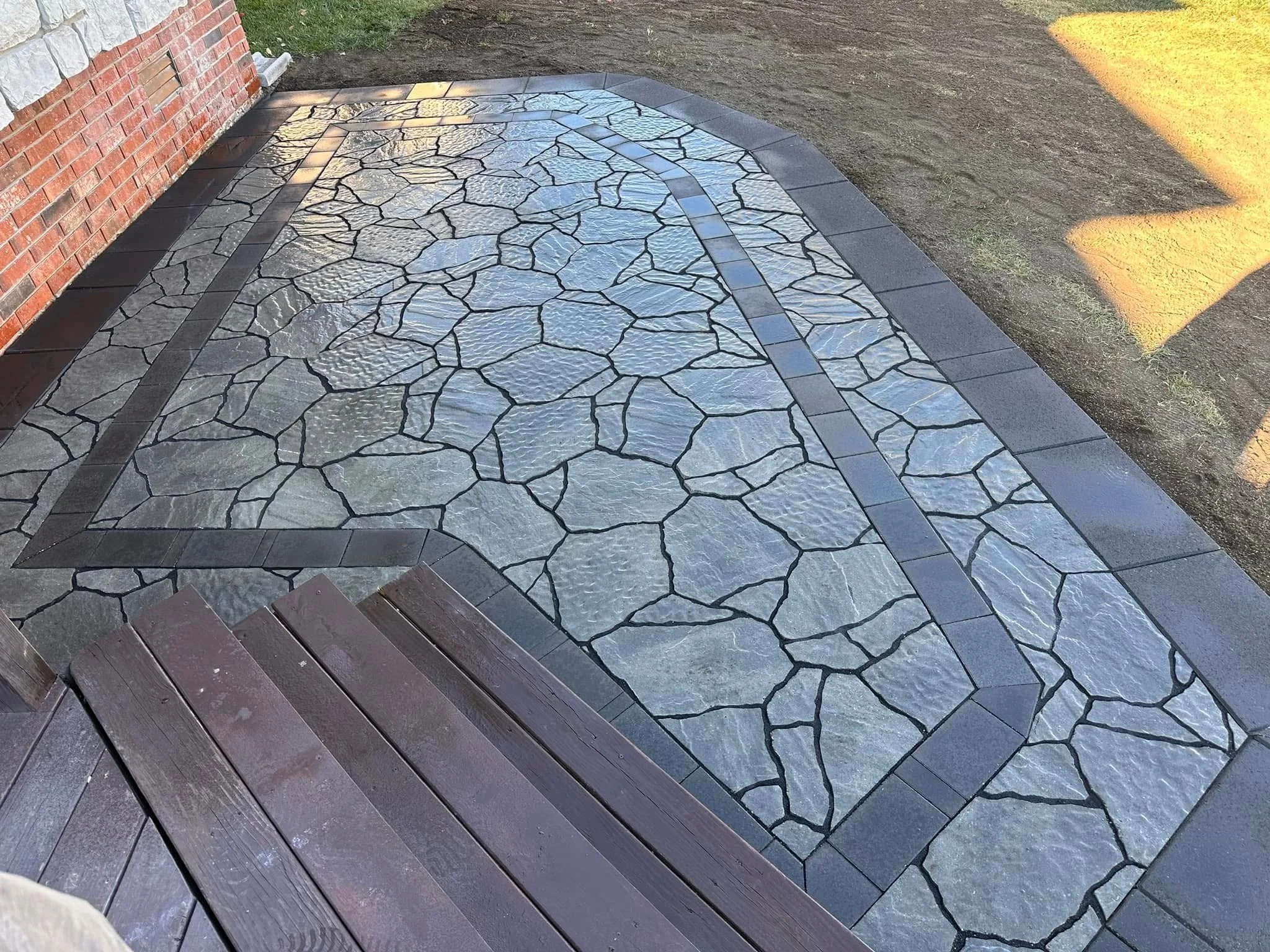 Decorative outdoor stone walkway bordered by dark gray bricks, adjacent to a brick house and a patch of soil with grass on the side.