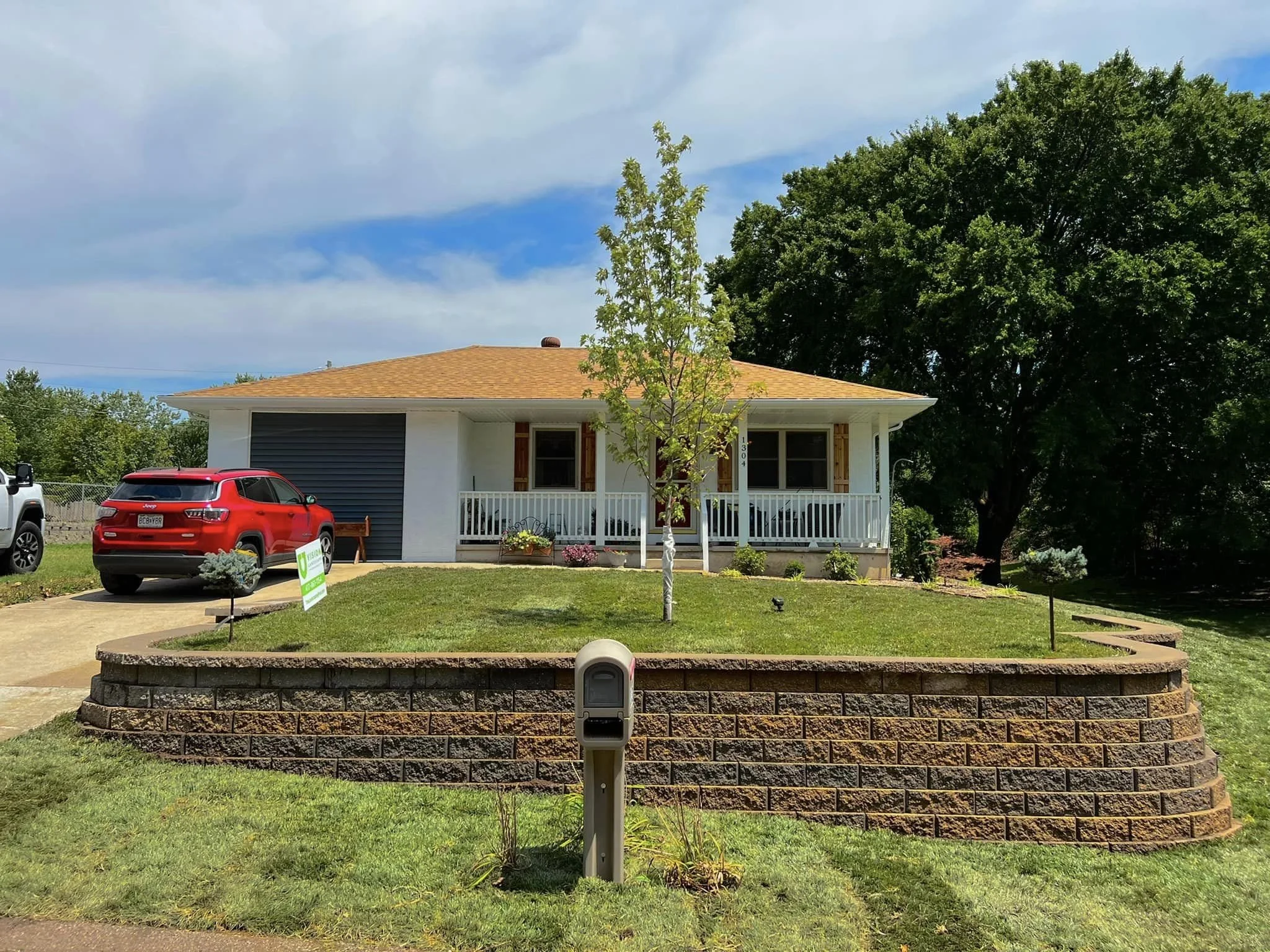 A house with a brown roof, white exterior, and small porch, surrounded by a green lawn, small trees, and flowering plants. Two cars are parked in the driveway, and there is a brick wall with a mailbox in the foreground. Large trees are in the backgro