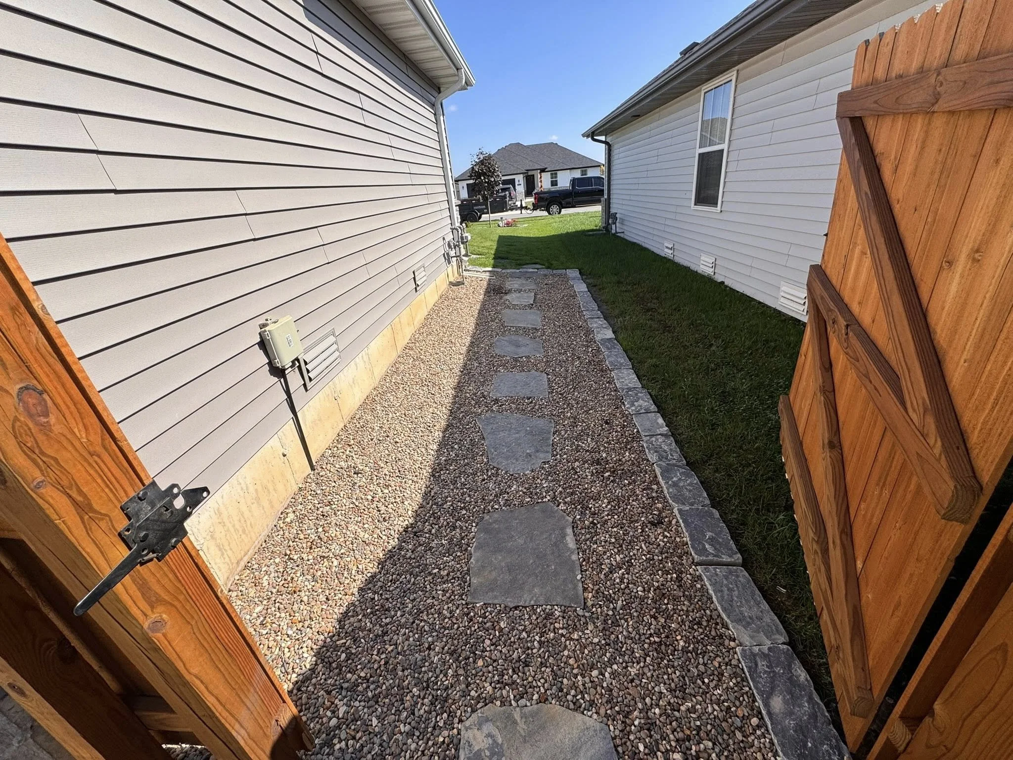 A backyard with a gravel pathway bordered by stones, a wooden fence, and two houses with vinyl siding, with a sunny sky in the background.