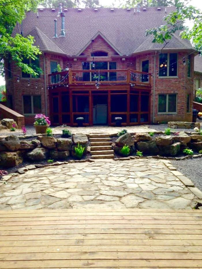 Backyard with stone pathway, wooden stairs, garden plants, and a brick house with a screened porch and upper deck.