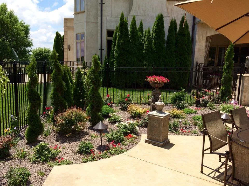 Beautiful backyard garden with tall evergreen trees, colorful flowers, a stone fountain, and a patio with chairs and a large umbrella.