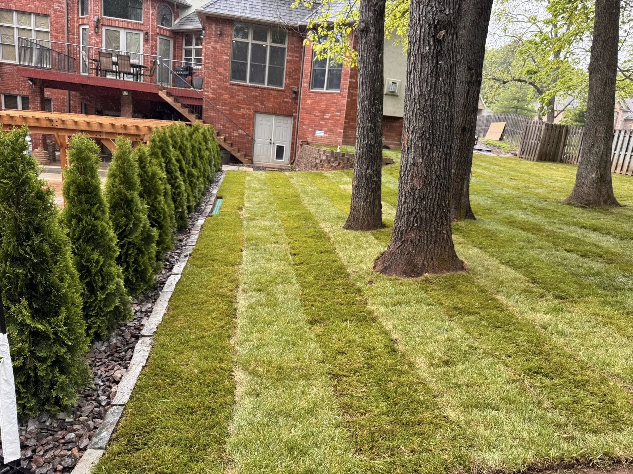 A backyard with a well-maintained lawn, three large trees, and a brick house with a deck. The deck has chairs and a grill, and is accessed by a staircase. A fence borders the yard.