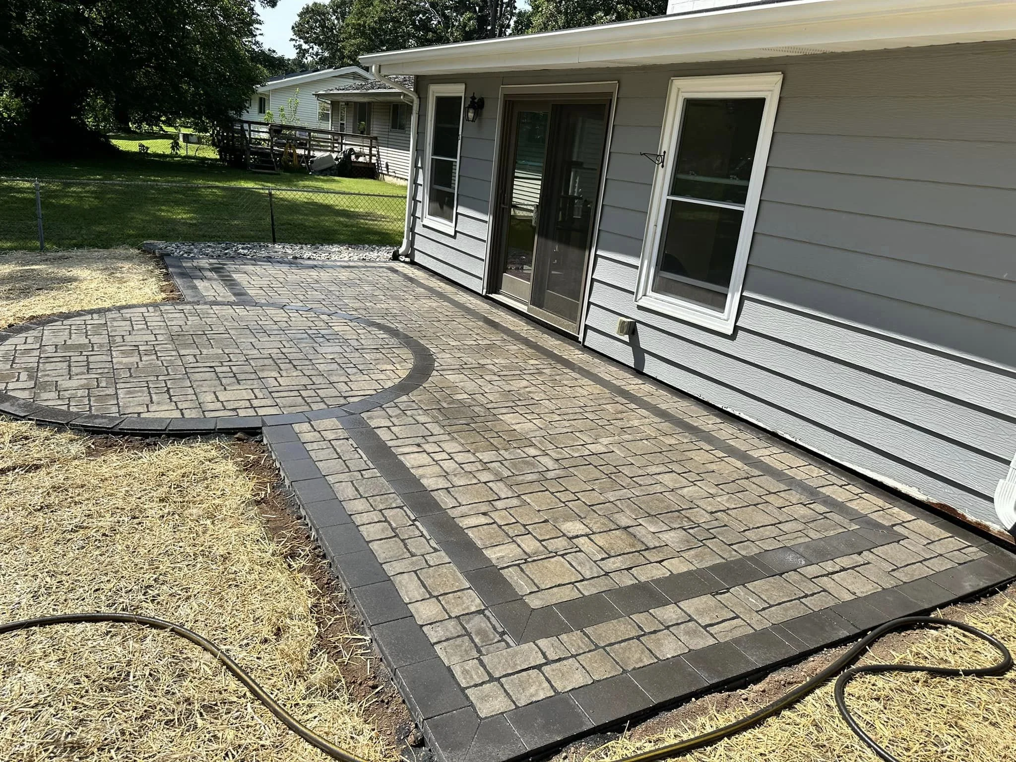 Backyard patio with brick and stone pavers, a sliding glass door, and a grey house. The yard has grass, a chain-link fence, trees, and a small deck in the background.