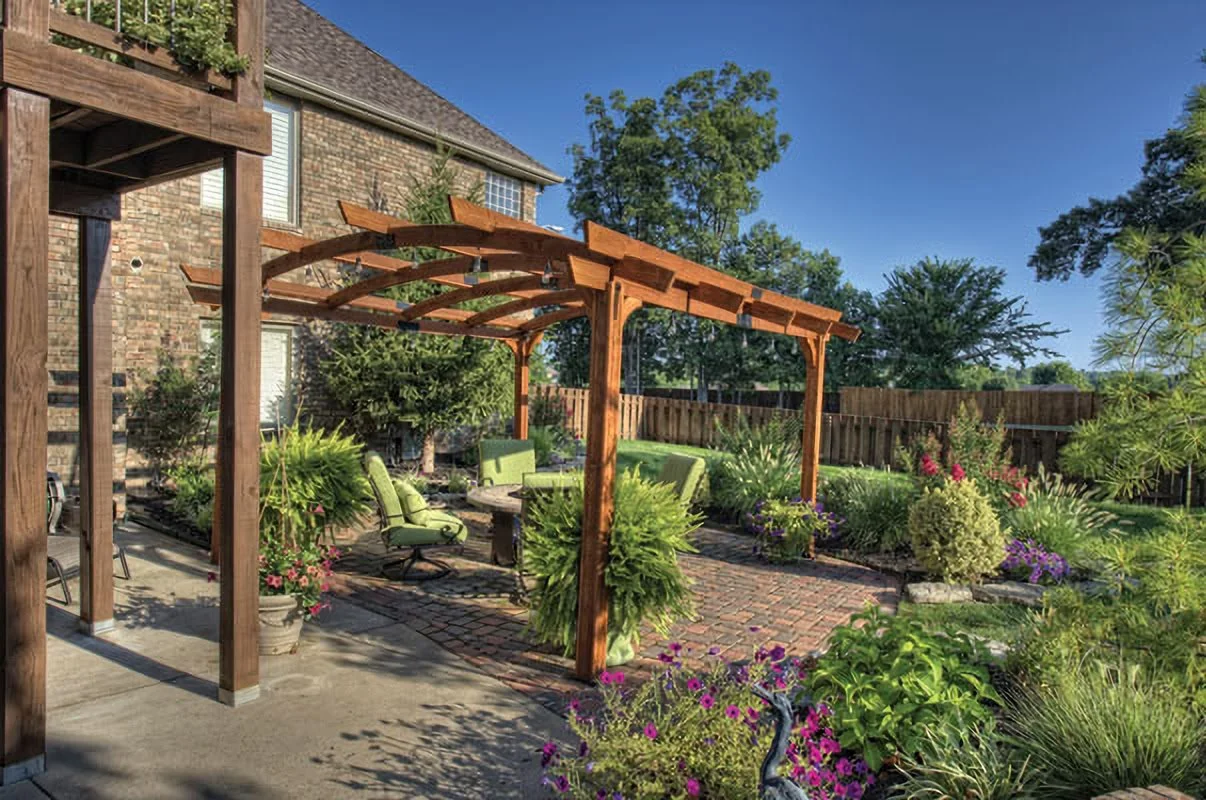 Backyard garden with a wooden pergola, green outdoor chairs, potted plants, and colorful flowers, enclosed by a wooden fence, on a sunny day.