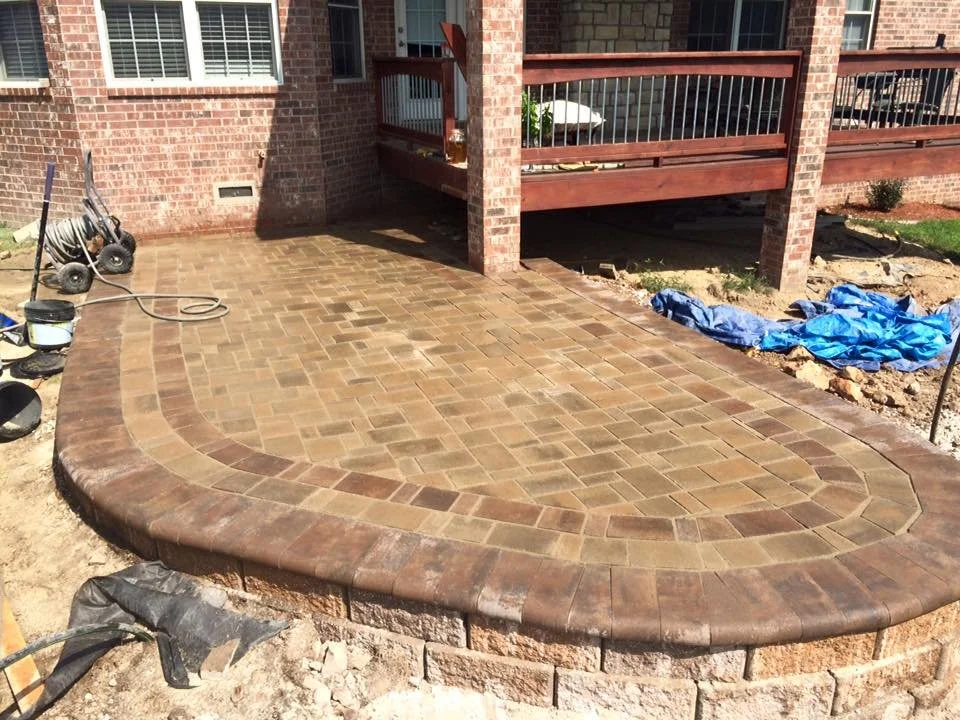 Newly constructed brick paver patio with curved edge, adjacent to a brick house with a wooden deck, construction tools, and blue tarp on the ground.