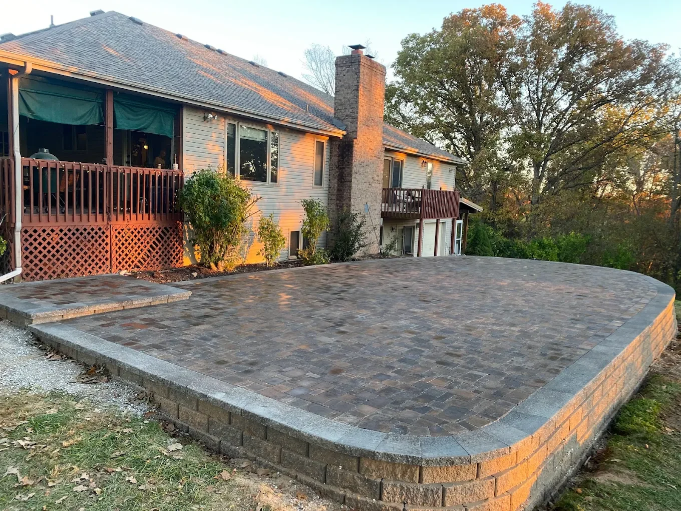Backyard with newly paved stone patio, house with balcony, and trees in the background