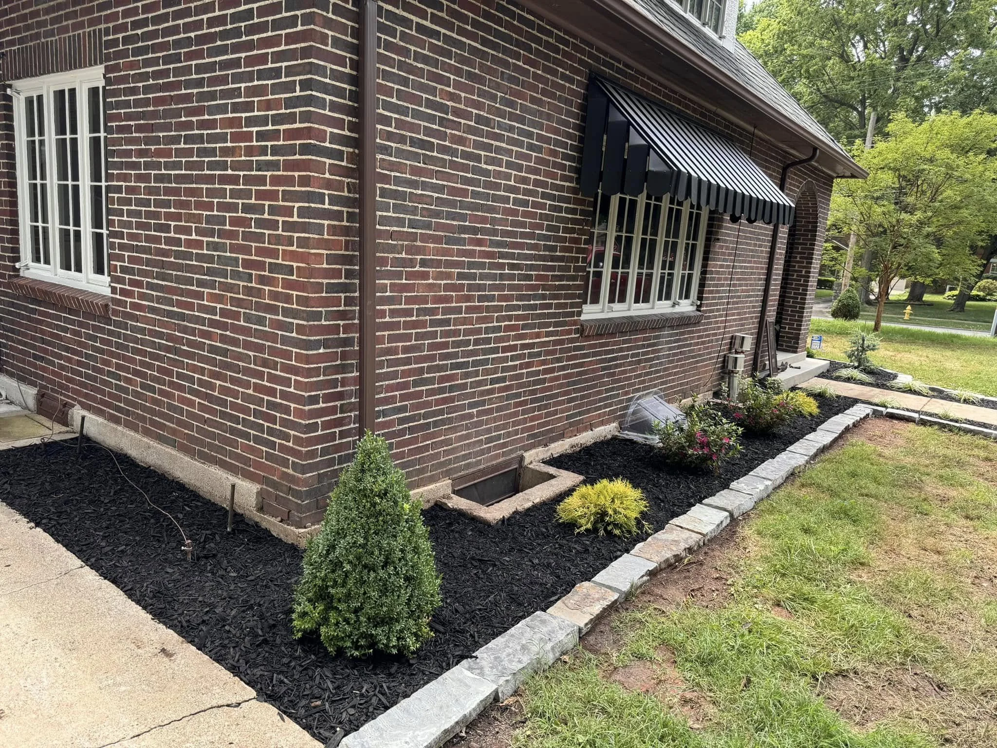 A brick house with white-framed windows and a striped black awning above one window. The house has a brown gutter and downspout. In front, there's a small garden with mulch, green shrubs, and colorful flowers, bordered by a stone edging. The lawn is 