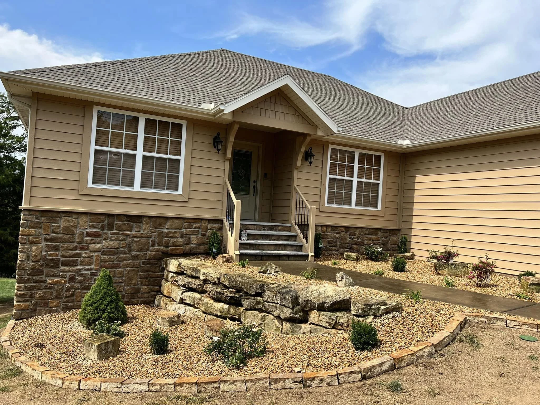 A beige house with stone foundation, front steps, and manicured front yard with bushes and small rock wall, under a blue sky with clouds.